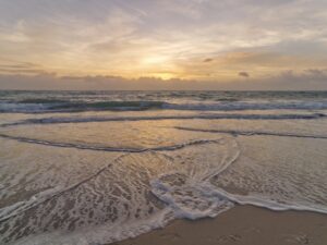 Gentle ocean waves reaching the shore at sunrise with soft light across the water