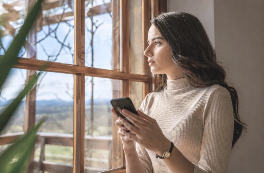 Woman looking out a window while holding her phone, reflecting after seeing a difficult story